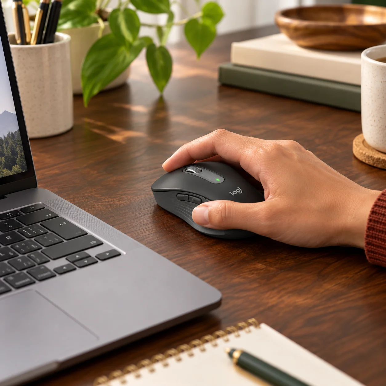 Hand using a graphite Logitech Signature M650 L wireless mouse beside a laptop on a wooden desk.
